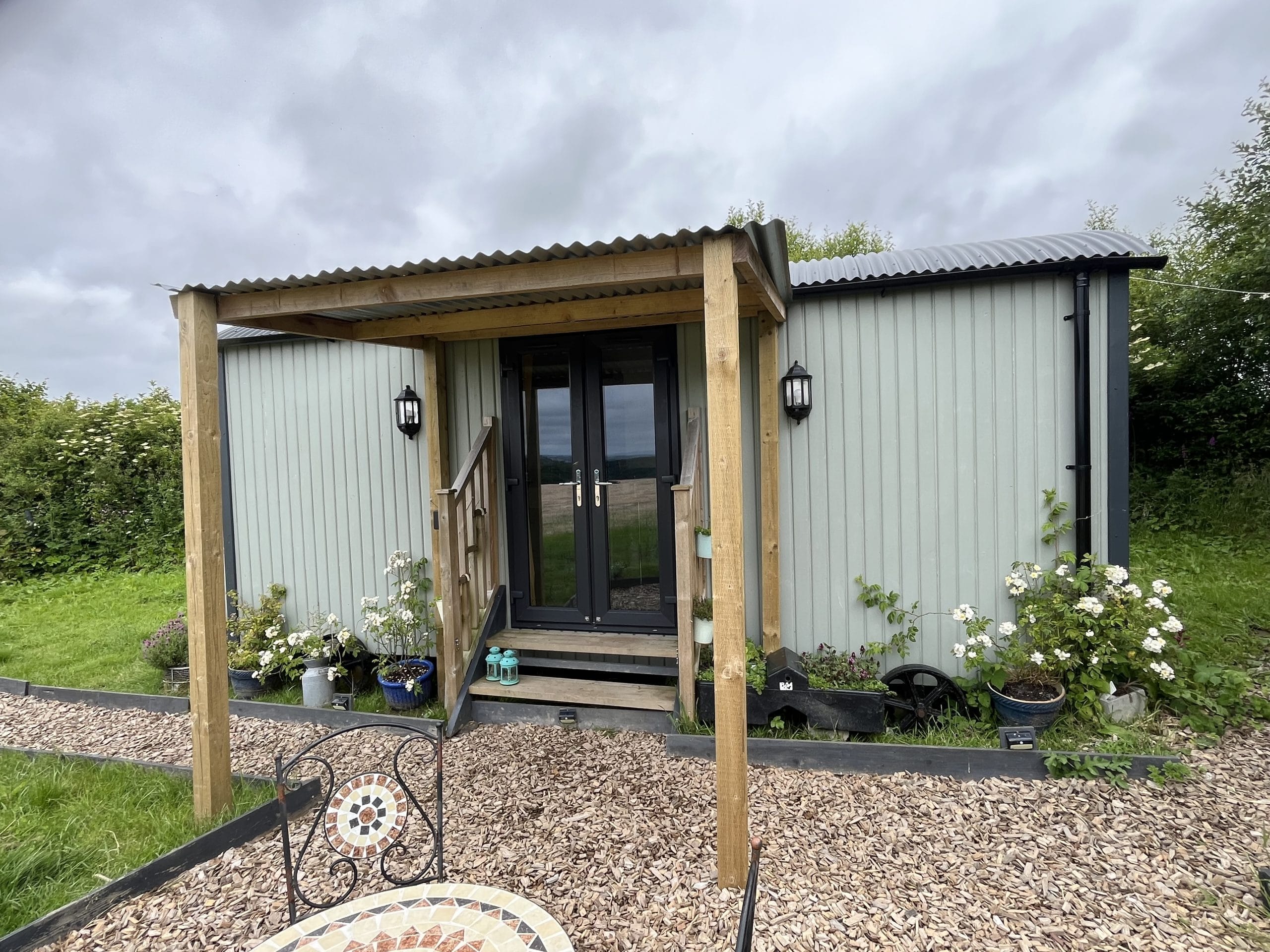 Entrance of a modern tiny house with wooden steps and plants, featuring a black door and gray metal siding.
